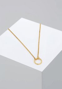 Collier délicat en chaîne d'or avec un pendentif circulaire lisse et poli reposant sur une surface géométrique blanche sur un fond gris.