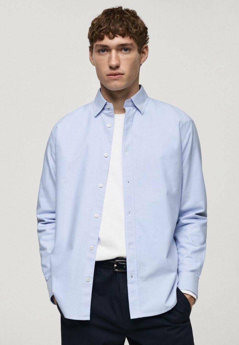Chemise boutons bleue claire avec un col étalé, manches longues et boutons blancs ; portée sur un t-shirt blanc, associée à un pantalon sombre.