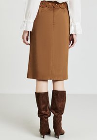 Gonna a matita in satin marrone con una cintura in pelle a scallop con fori circolari. Indossata con stivali alti in suede marrone a collo basso.