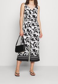 Mono floral en blanco y negro con escote en V, piernas acampanadas anchas y un bolso negro con un patrón texturizado. Se muestran chanclas.