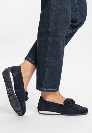 Navy suede loafers with a bow accent, rubber sole, and contrasting white trim. Worn with dark denim trousers rolled at the cuff.