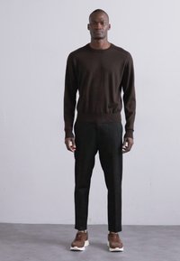 Hnědý pletený svetr s kulatým výstřihem, doplněný černými konfekčními kalhotami a hnědými teniskami s bílou podešví. Modelka stojí na neutrálním pozadí.