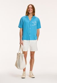 Lysblå skjorte med korte ærmer og hvide broderede grafik, parret med hvide shorts og beige høje sneakers, mens han/hun holder en beige tote bag.