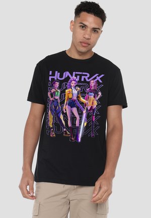 Young man wearing black T-shirt featuring three stylized female characters in futuristic outfits with purple "HUNTRX" text background.