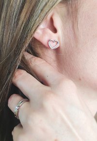 Orecchini argentati a forma di cuore adornati con piccole pietre trasparenti, indossati all'orecchio di una persona, con una mano che tocca delicatamente l'orecchio.
