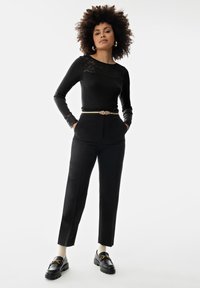 Zwarte lange mouwen top met kantdetail, gecombineerd met zwarte op maat gemaakte broeken, een gouden accentriem en chunky zwarte loafers over witte sokken.