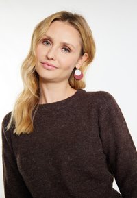 Brązowy sweter dziergany z okrągłym dekoltem, w połączeniu z czerwonymi okrągłymi kolczykami z teksturowanym srebrnym akcentem. Gładkie fale włosów.