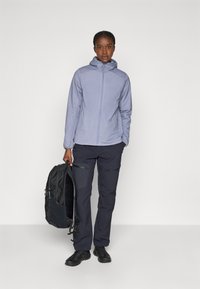 Lyselilla zip-up jakke med hette, paret med mørke bukser og svarte sko. Figuren holder en svart ryggsekk og står foran en ensfarget bakgrunn.