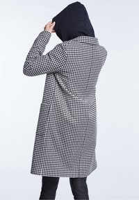 Persona vestida con un abrigo de pata de gallo en blanco y negro y sudadera con capucha negra, ajustando la capucha, de espaldas frente a un fondo claro y liso.