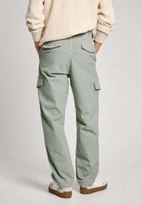 Svetlozeleni cargo hlače iz mešanice bombaža, z bočnimi žepi in sproščenim krojem, v kombinaciji z belimi športnimi copati na nevtralni podlagi.