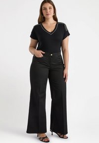 Schwarzes V-Ausschnitt-T-Shirt mit verzierten Schulterdetails, kombiniert mit hoch taillierten schwarzen weiten Hosen. Die Person steht in schwarzen High Heels.
