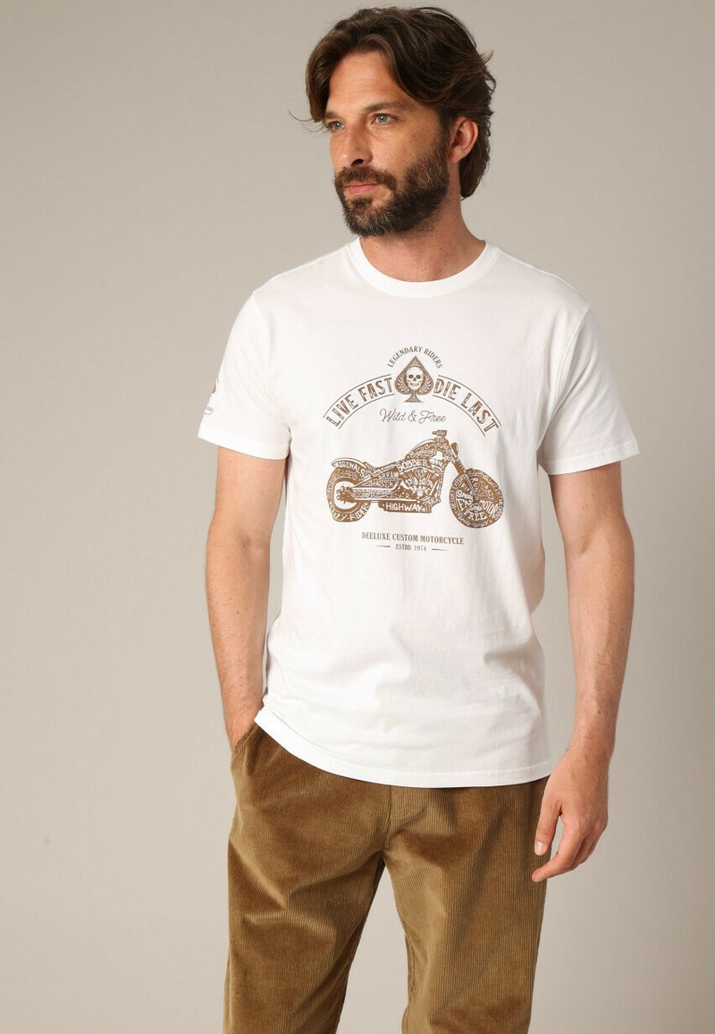 T-shirt en coton blanc avec un imprimé graphique marron représentant une moto et le texte "Vivez vite, mourrez dernier", assorti à un pantalon en velours côtelé marron.