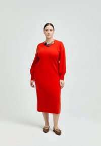 Rotes Wollkleid mit langen Ballonärmeln, knielang, runder Ausschnitt. Kombiniert mit braunen Ballerinas, akzentuiert durch eine mehrfarbige Halskette.
