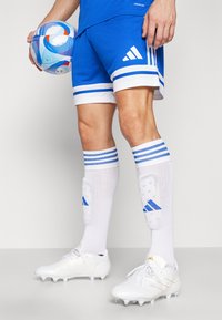 Blå fotbollsshorts med vita ränder, vita fotbollsstrumpor med blå detaljer och vita fotbollsskor med guld detaljer, håller en färgglad fotboll.