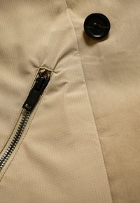 Bézs színű szövet hálómintával, fekete gombbal és fekete cipzárral. Az anyag sima textúrájú, enyhén fényes felülettel.