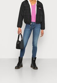 Czarna ocieplana kurtka z białym polarem, różowy sweter z dekoltem w serek, niebieskie rurki i czarne buty wojskowe, trzymająca czarną torebkę.