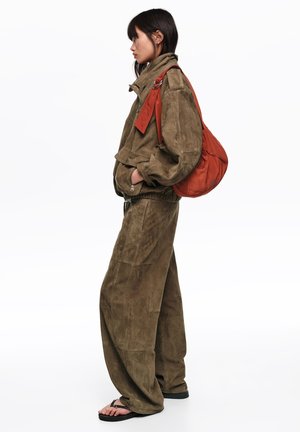 Mujer de pie con chaqueta de ante verde oliva y pantalones anchos, manos en los bolsillos, llevando un gran bolso de hombro color óxido, con sandalias negras.