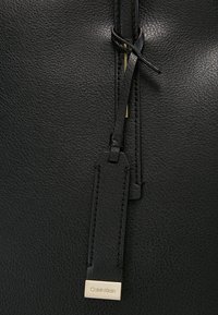 Black textured leather handbag with flat strap and gold-tone hardware. Features a branding tag with engraved "Calvin Klein" logo.