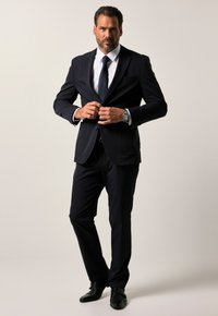 Completo blazer navy e pantaloni, vestibilità sartoriale, tessuto liscio, camicia bianca, cravatta scura e scarpe eleganti nere, con orologio d'argento al polso.