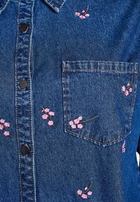 Camisa de denim con cuello, botones negros y bordado floral rosa en el pecho y en el bolsillo. Cuenta con un solo bolsillo frontal.