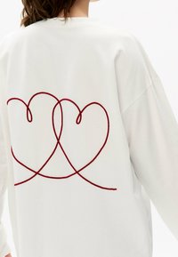 Weißer Pullover mit einem roten, gestickten Design aus ineinander verschlungenen Herzen auf der Rückseite. Weicher Stoff mit entspannter Passform und überschnittenen Schultern.