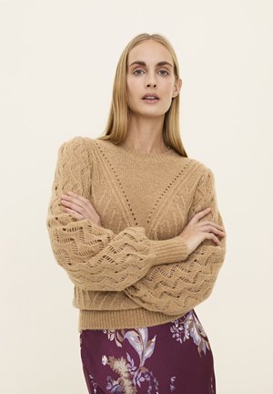 Knit sweater in light brown with a textured pattern, featuring voluminous sleeves and a ribbed hem. Paired with a floral skirt.