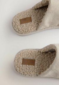 Beige Hausschuhe mit einem Obermaterial aus strukturiertem Stoff, weicher, flauschiger Innenausstattung und einem kleinen Lederpatch an der Sohle. Rundliche Zehenform, ohne Absatz.