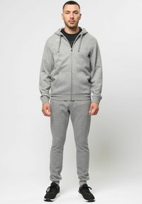 Felpa grigia con zip e cordini, dotata di tasche frontali, abbinata a pantaloni sportivi grigi coordinati. Sneakers nere completano l'outfit.