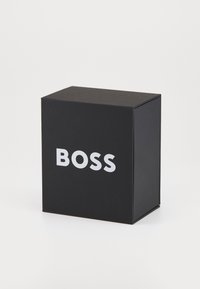 Melnas taisnstūra kaste ar matētu apdari, kuras priekšpusē ir vārds "BOSS" resnā baltā fontā. Vienkāršs un kompakts dizains.