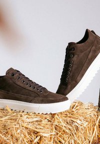 Braune Wildleder-Hochschaft-Sneaker mit weißer Gummisohle, ausgestattet mit runden Metallösen und flachen Schnürsenkeln, auf einem Strohuntergrund platziert.