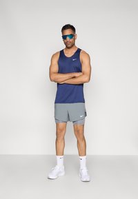Maiou de performanță Nike bleumarin, asortat cu pantaloni scurți gri. Modelul poartă ochelari de soare sportivi, adidași albi și șosete cu logo Nike.