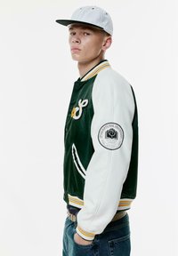Groene en witte varsityjack met gele accenten, voorzien van een geborduurde bloem, gestreepte ribbelkraag en een logo-opdruk op de mouw.