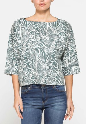 Donna che indossa una maglia larga bianca con motivo di foglie verdi, maniche a tre quarti e jeans blu su uno sfondo semplice.