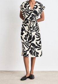 Robe enroulée à motif feuille en noir et blanc avec des manches courtes, un décolleté en V, une ceinture à la taille et une longueur sous le genou. Texture de tissu lisse.