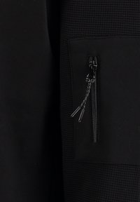 Chaqueta negra con tejido texturizado, que cuenta con un bolsillo con cremallera en la parte delantera y tiradores de cremallera. Diseño elegante y moderno.