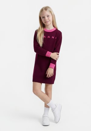 Junges Mädchen steht mit gekreuzten Beinen und trägt ein bordeauxrotes Strickkleid mit rosa Bündchen und Kragen, weiße Socken und weiße Turnschuhe.