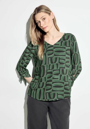 Blouse verte imprimée avec un décolleté en V, présentant un motif géométrique audacieux en vert foncé et noir. Manches longues avec des accents noués aux poignets.