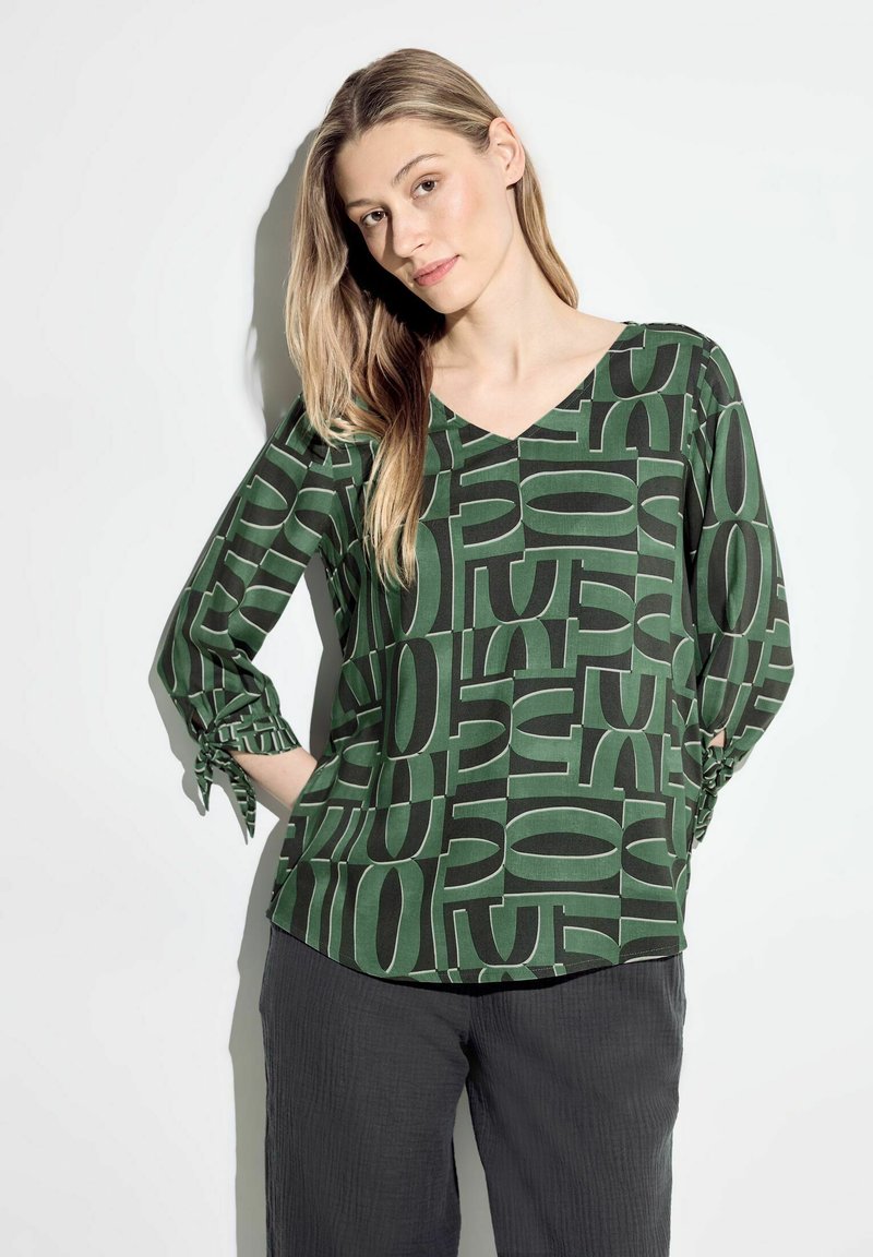 Blusa verde stampata con scollatura a V, caratterizzata da un audace motivo geometrico in verde scuro e nero. Maniche lunghe con dettagli legati ai polsini.