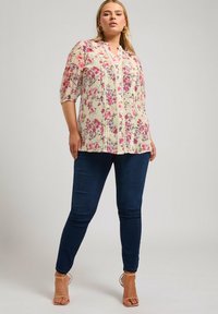 Kukkakuvioinen tunika vaaleassa kermavärissä, jossa on vaaleanpunaisia ja purppuran värisiä kukkia. Tunicassa on nappikiinnitys edessä, pyöreä kaula-aukko ja kolmanneksen pituiset hihat, ja se on yhdistetty tummiin farkkuihin.