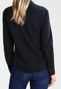 Femme aux longs cheveux blonds portant une chemise noire ajustée à boutons et un jean bleu foncé, vue de dos sur un fond uni.