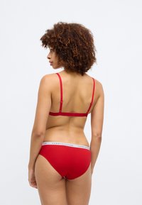 Punane pesu komplekt, mis sisaldab silmapaistmatut rinnahoidjat õhukeste rihmadega ja sobivaid aluspükse bränditud elastse vöökohaga.