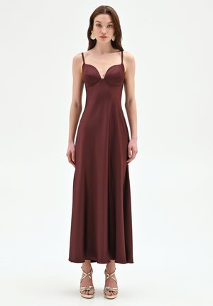 Donna che indossa un abito bordeaux lungo fino al pavimento, senza maniche, con sottili spalline e sandali dorati con tacco alto, in piedi davanti a uno sfondo semplice.