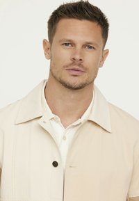 Beige colbert met knoopsluiting, gemaakt van een soepel materiaal. Draagt over een wit shirt, met een strakke en minimalistische uitstraling.
