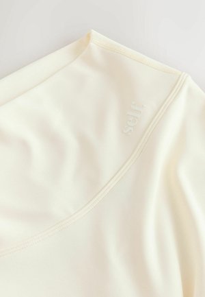 Top crem realizat dintr-un material neted și elastic, cu un decolteu larg și un logo discret în relief. Finisajul include cusături vizibile.