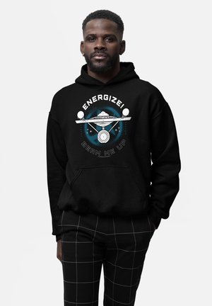Mężczyzna w czarnej bluzie z kapturem z grafiką statku kosmicznego i napisem "ENERGIZE! BEAM ME UP," w połączeniu z czarnymi spodniami z białymi liniami w kratkę.