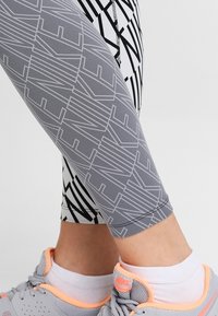 Leggings atletici grigi con un motivo geometrico, caratterizzati da un design aderente e una texture liscia. Indossati con scarpe grigie e accenti arancioni.