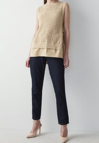 Beige stickad ärmlös topp med texturerat mönster över en ljusare bas, kombinerad med mörka denimjeans och nude högklackade skor.