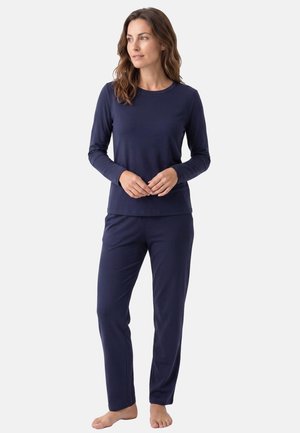 Femme debout pieds nus portant un haut bleu marine à manches longues et un pantalon assorti, regardant légèrement sur le côté devant un fond uni.