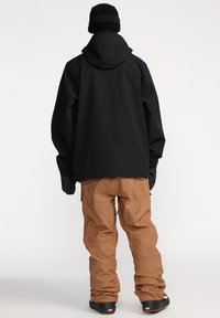 Veste noire avec capuche et coupe ample, associée à un pantalon de neige marron, fuselé. La tenue présente un tissu texturé et des poignets élastiques.
