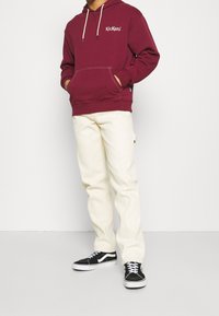 Person, der einen kastanienbraunen Kickers-Pulli, cremefarbene Hose und schwarz-weiße Sneakers trägt, steht mit den Händen in der Hoodie-Tasche.